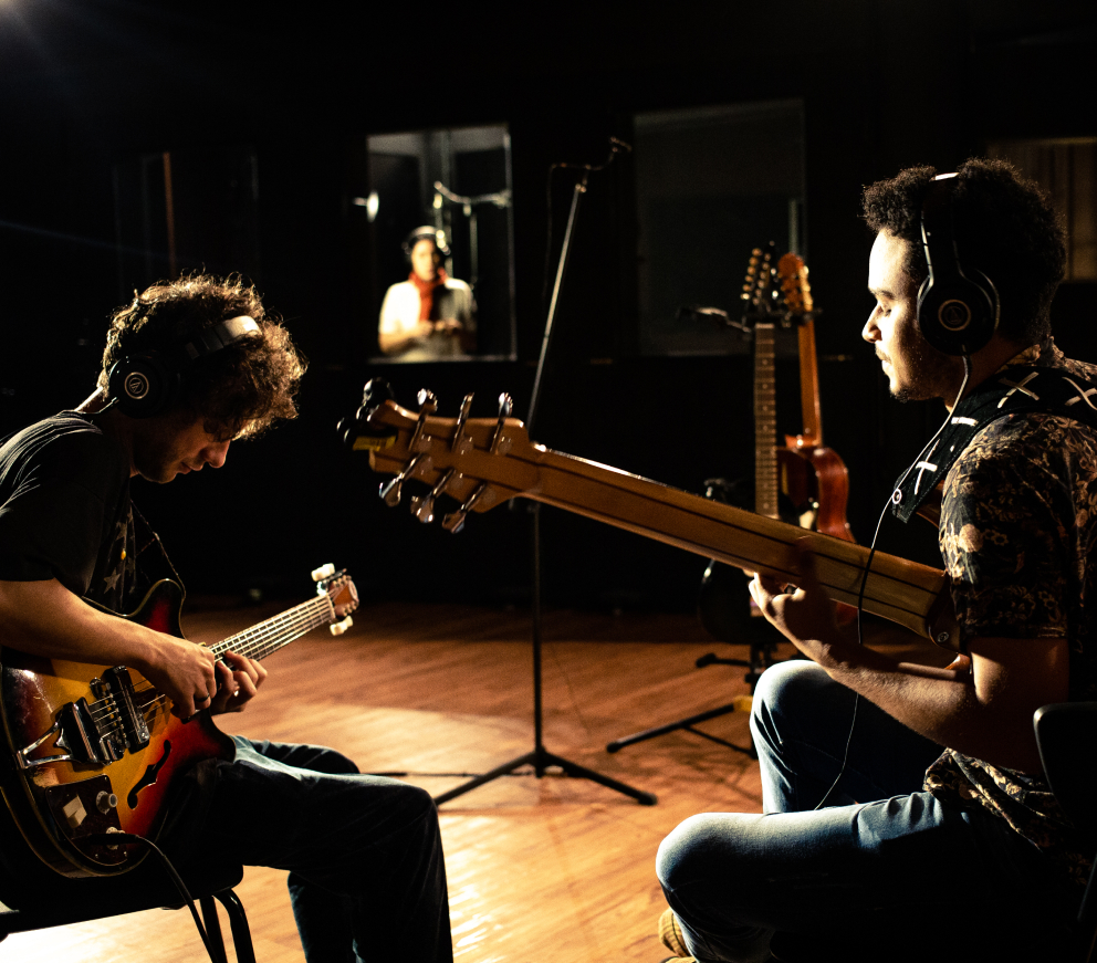 Dois homens tocam guitarra enquanto uma mulher canta em um estúdio de gravação, criando uma atmosfera musical vibrante.