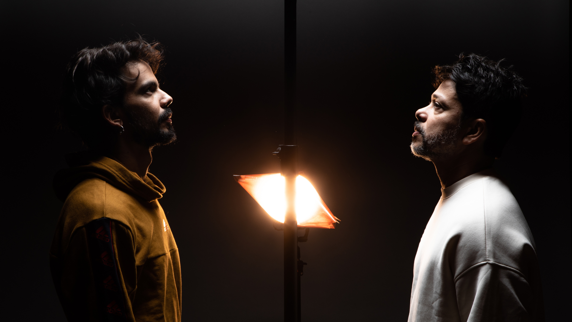 Dois homens se posicionam em frente a uma fonte de luz, destacando suas silhuetas em um cenário com baixa luz.